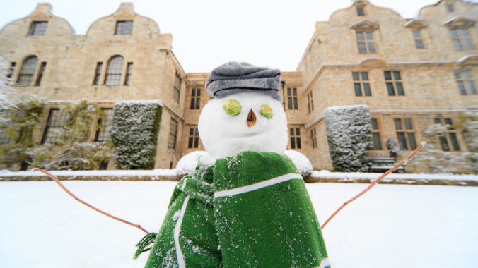 Snowman at the front with a snowy garden and stone house behind